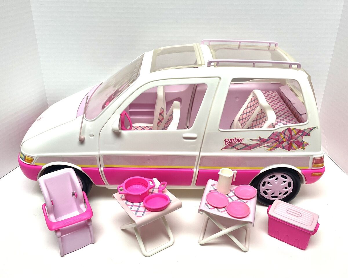 A white and pink Barbie minivan beside a pink car seat, fold tables, dishes, and cooler